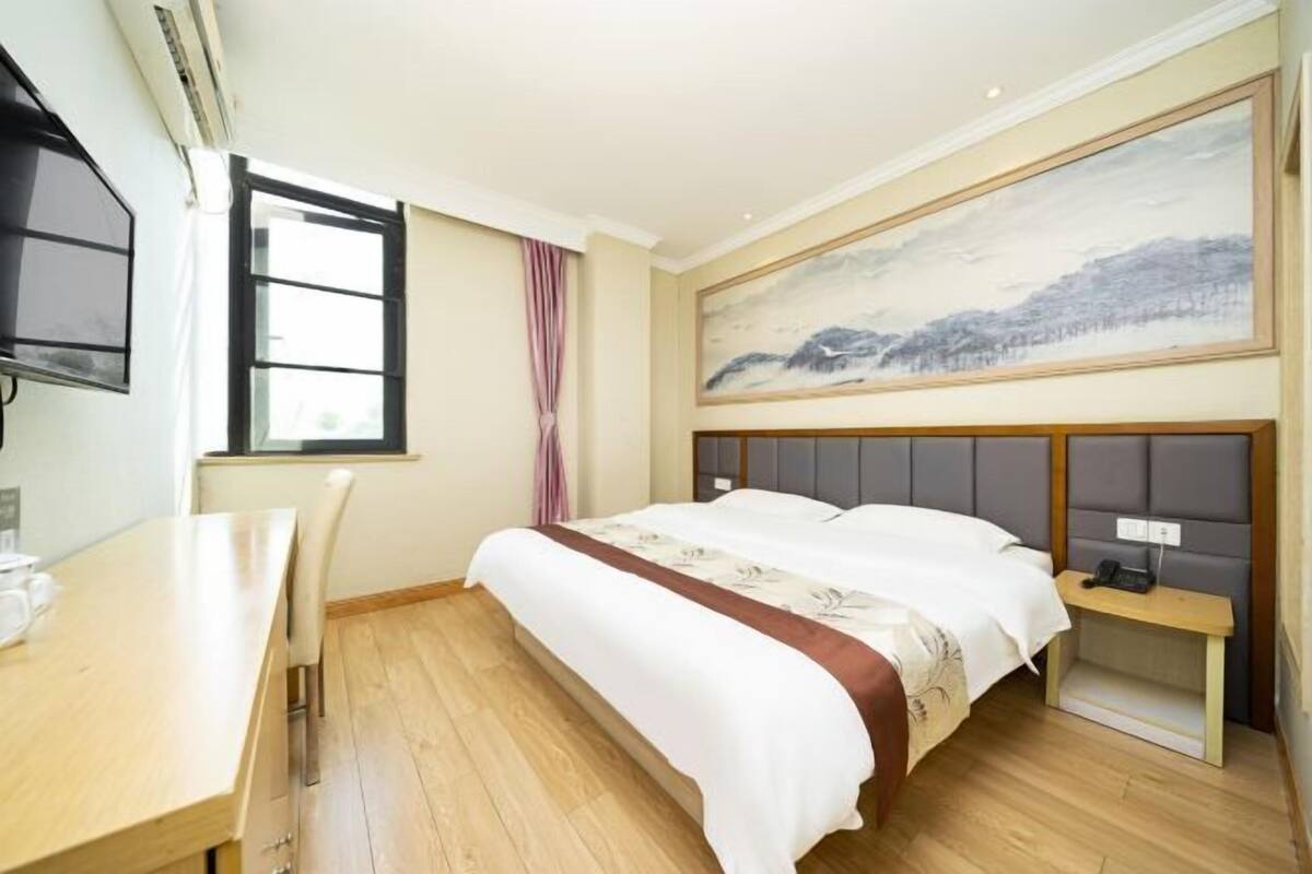 Habitación de hotel con cama, escritorio, silla, ventana, un cuadro en la pared y una mesita de noche con lámpara.