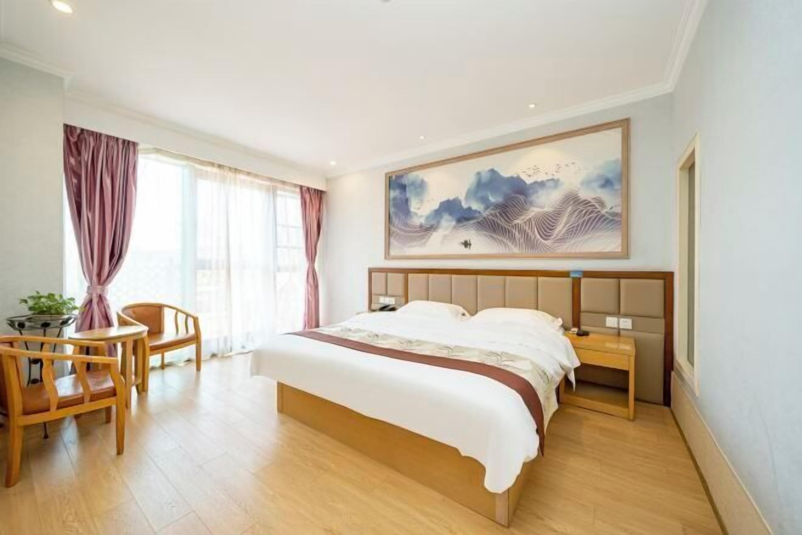 Ein Hotelzimmer mit einem großen Bett, einem Holz-Kopfstück, einem Nachttisch, einem Sessel, einem Schreibtisch und einem großen Gemälde an der Wand.