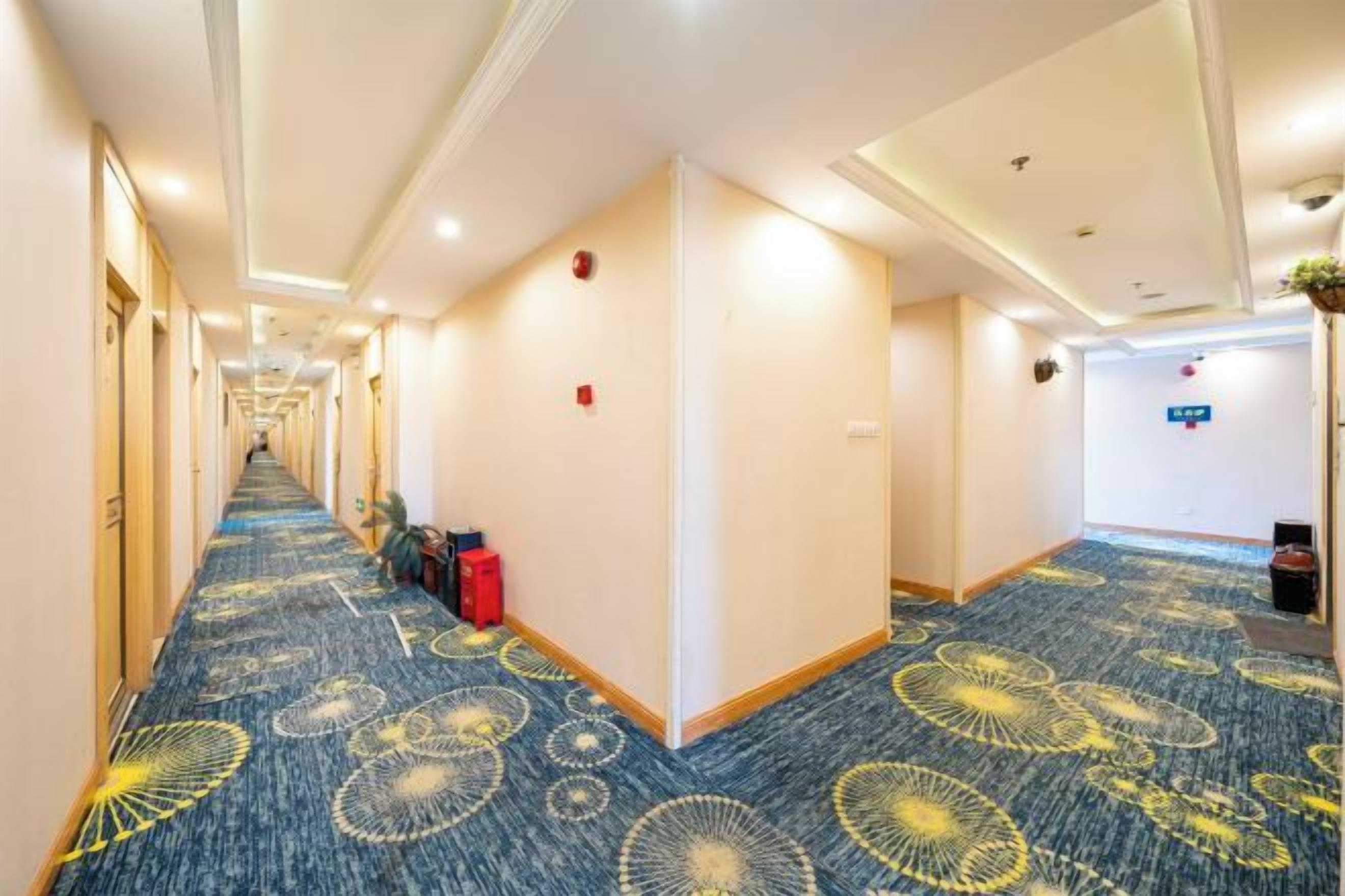 A hotel corridor with a patterned carpet, doors on both sides, and a reception area in the distance.