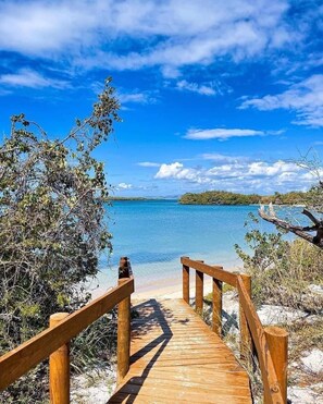Pasarela de madera que lleva a una playa con agua azul cristalina y una isla a lo lejos.