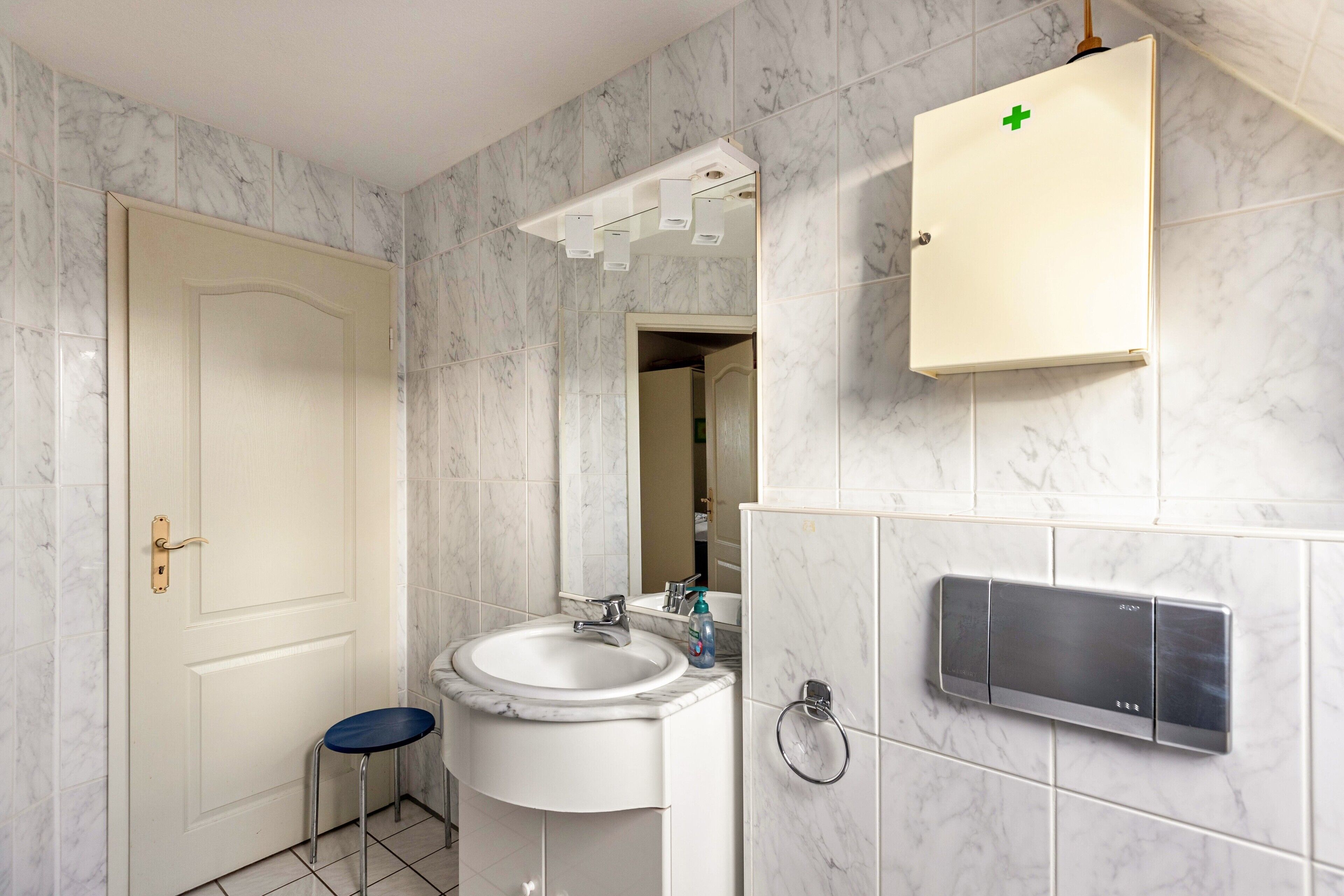 A bathroom with a white sink, a mirror, a cabinet with a green cross, and a small stool.