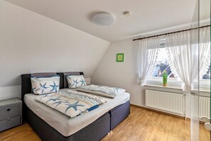 Une chambre à coucher avec un lit, une table de chevet, une fenêtre avec des rideaux, un radiateur et un petit tableau accroché au mur.