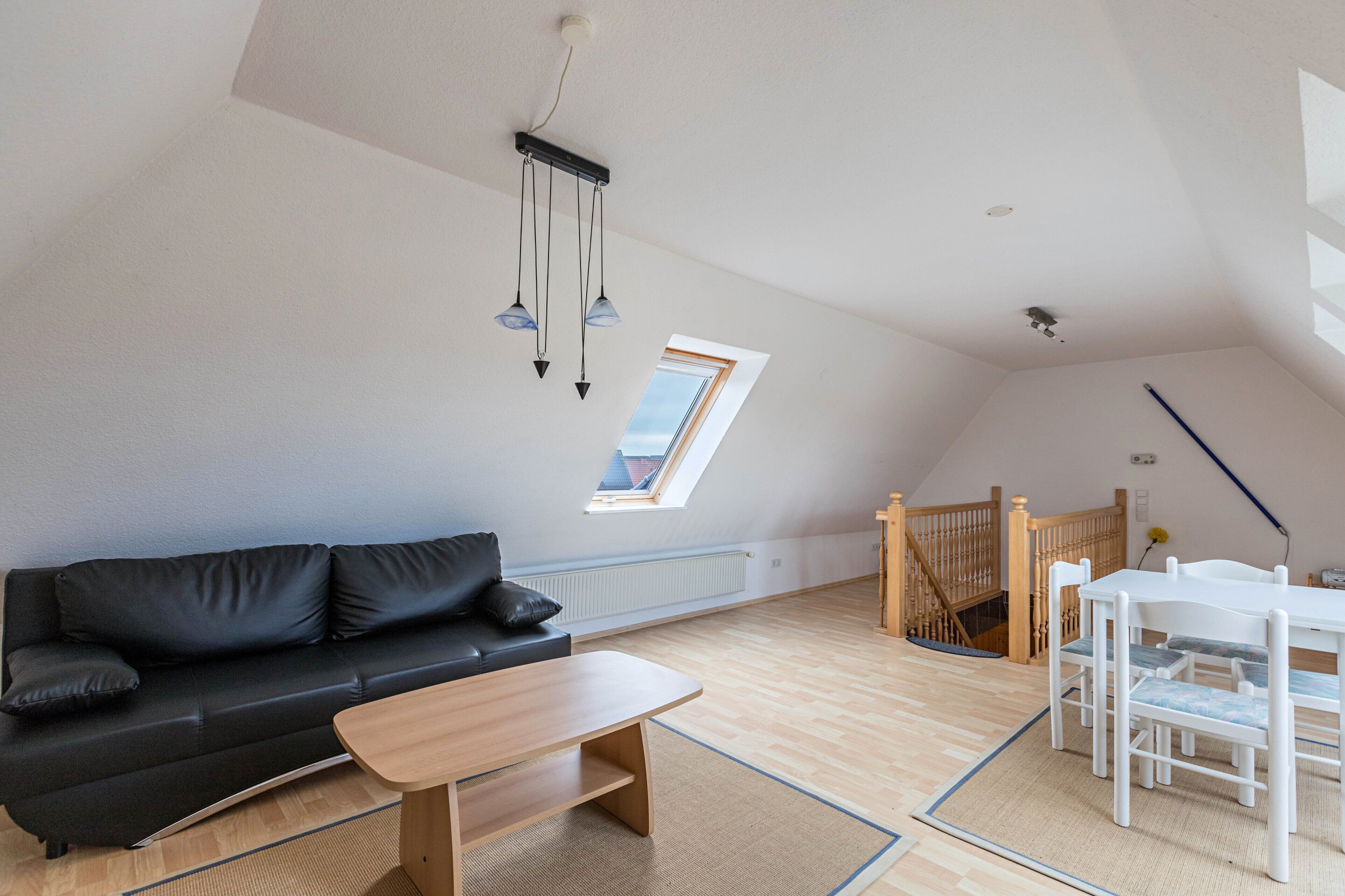 Une pièce comprenant un canapé en cuir noir, une table en bois, un ensemble de salle à manger blanc, une lucarne et un escalier en bois.