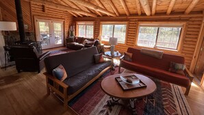 A log cabin interior with a wooden ceiling, a fireplace, a sofa, a red couch, and a dining table with a tray. - Cozy cabin in the woods. 12 min from the Angel Fire ski resort; 6 to golf course (Angel Fire)