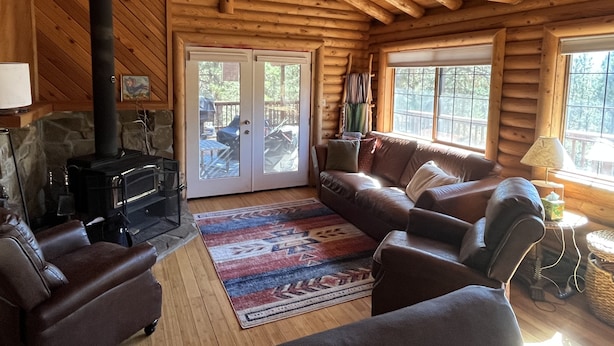 A log cabin interior with a fireplace, leather sofa, and a large window. - Cozy cabin in the woods. 12 min from the Angel Fire ski resort; 6 to golf course (Angel Fire)