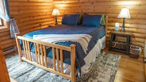 A log cabin bedroom with a wooden bed, two bedside lamps, a small table, and a window with curtains. - Cozy cabin in the woods. 12 min from the Angel Fire ski resort; 6 to golf course (Angel Fire)