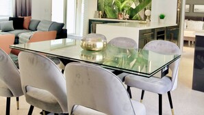 Een moderne eethoek met een glazen tafel en grijze stoelen met bekleding, een centraal eiland met een plant, en een moderne hanglamp.