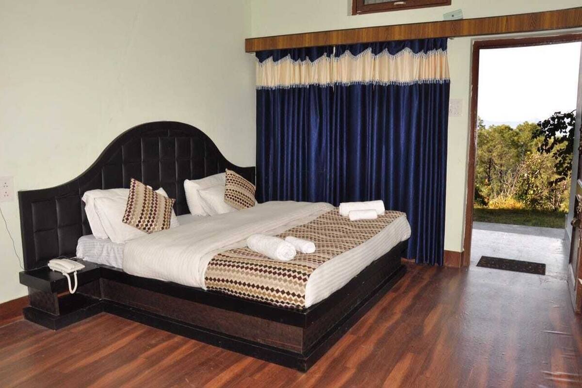 A neatly made bed with a patterned bedspread, a bedside phone, and a view of the outdoors through a window with blue curtains.