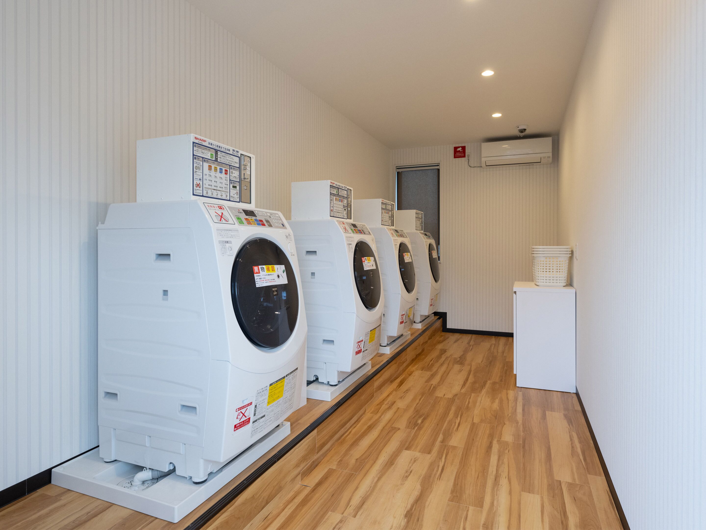 A row of white front-loading washing machines in a laundry room with wooden flooring and white walls.