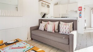 A modern living room with a grey sofa, a coffee table with a board game, a kitchen area with cabinets and a window, and a fire extinguisher on the wall. - Boutique style beach hut ideal for romantic getaways for all occasions. (Hele Bay, Ilfracombe)