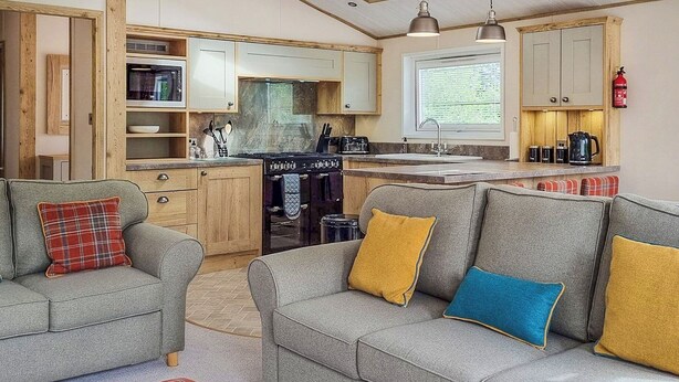 A compact kitchen with a stove, sink, and wooden cabinets. There are two grey sofas with colorful cushions and a plaid rug. - 1 bedroom accommodation in Woodhall Spa (Woodhall Spa)
