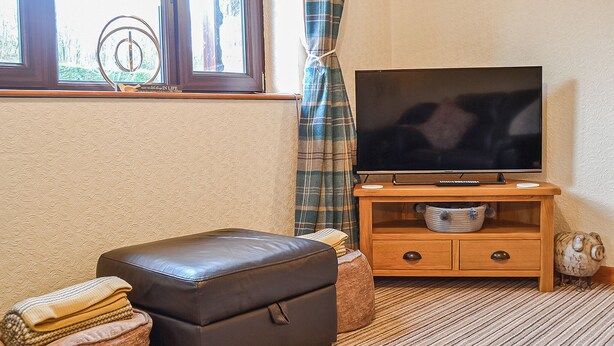 A room with a television on a wooden stand, a brown leather ottoman, a small wooden cabinet, and a window with curtains. - 1 bedroom accommodation in Dolgellau (Dolgellau)