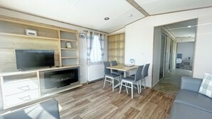 Ein modernes Wohnzimmer mit Flachbildfernseher, Holzregalen, einem Esstisch mit Stühlen und einem grauen Sofa.