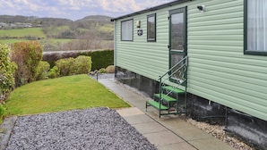 A green mobile home with a small porch, a green bench, and a view of the countryside.