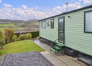 Uma casa móvel verde com um pequeno alpendre, um banco verde e vista para o campo.
