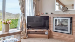 A modern living room with a flat-screen TV, fireplace, large windows with curtains, and a view of the outdoors.
