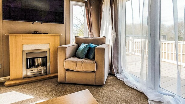Ein Wohnzimmer mit Kamin, einer Couch mit Kissen, einem an der Wand montierten Fernseher und großen Fenstern mit Vorhängen.