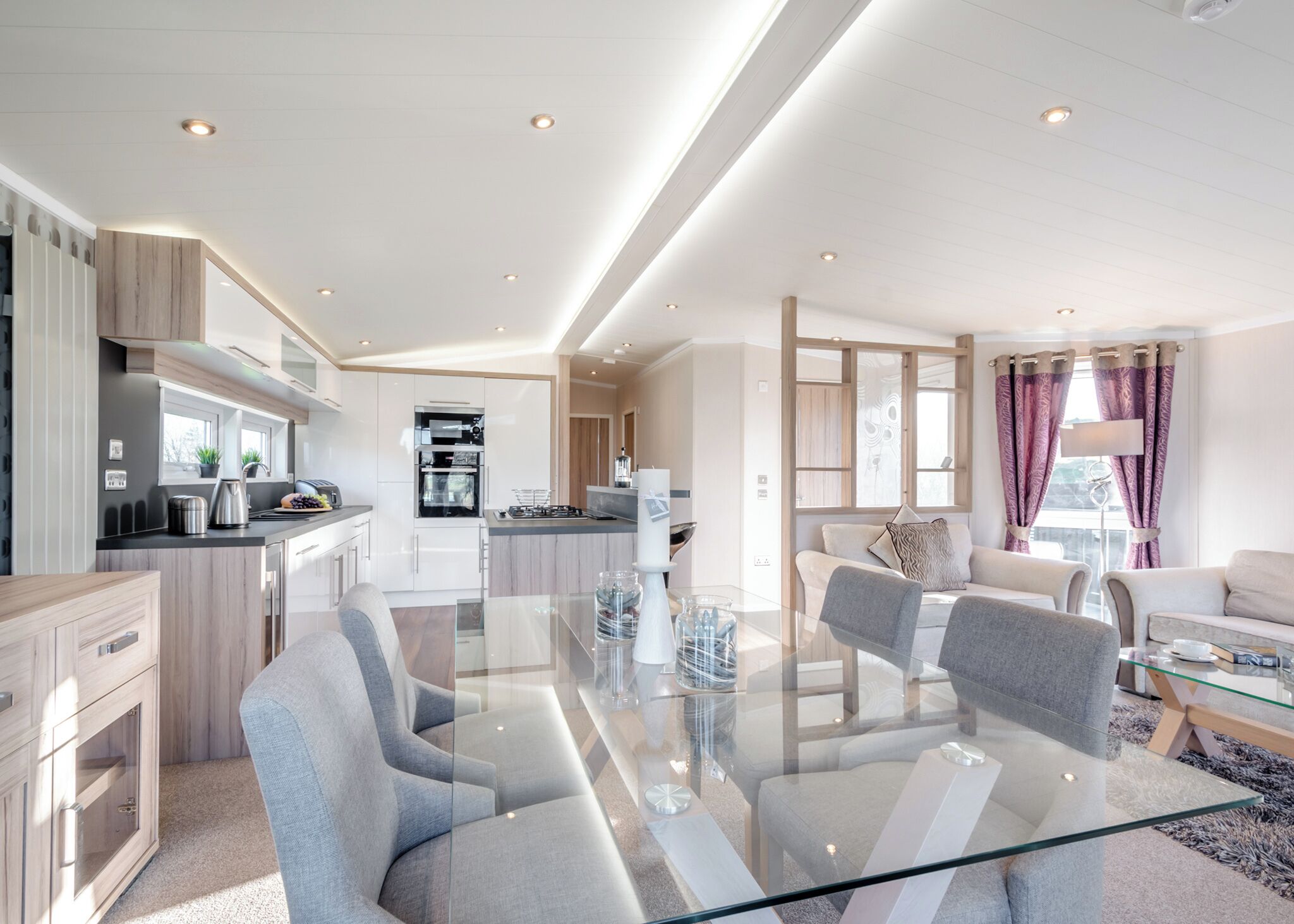 A modern kitchen with built-in appliances, a dining area with a glass table, and a living room with a sofa and a coffee table.