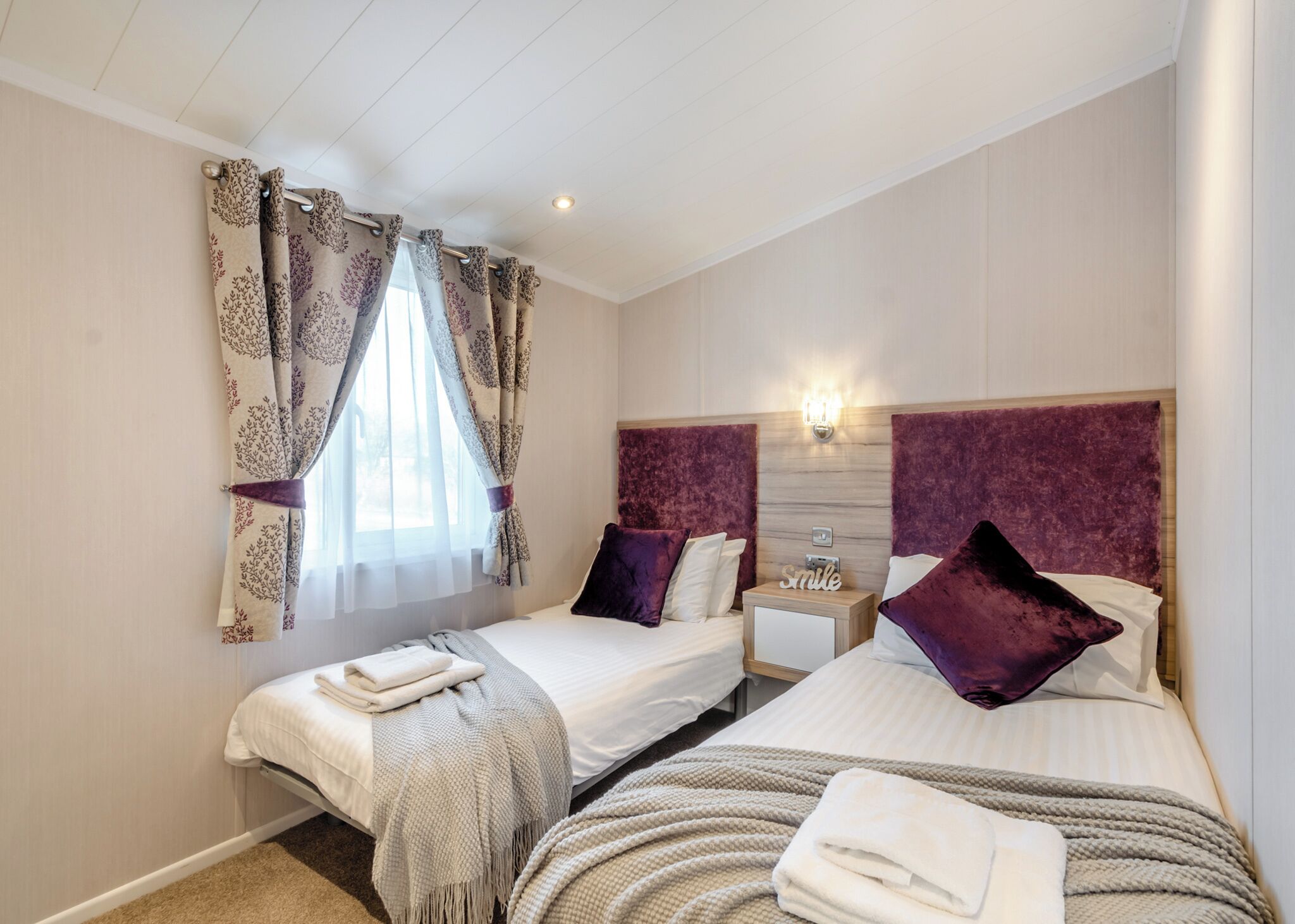 A hotel room with two beds, a window with curtains, a bedside table with a lamp, and a wall-mounted light fixture.