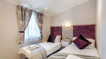 A hotel room with two beds, a window with curtains, a bedside table with a lamp, and a wall-mounted light fixture.