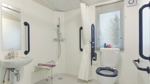 Une salle de bain avec un lavabo blanc, des toilettes, une douche avec un pommeau de douche amovible et une fenêtre avec des stores.