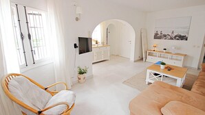 Una sala de estar luminosa con una silla de mimbre, un sofá color beige, un televisor y un comedor con mesa y sillas.