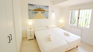 Habitación de hotel con cama, mesita de noche, un cuadro de una isla tropical y una ventana con persianas.