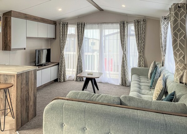 A modern living room with a sofa, a TV, a small table, and a balcony with curtains. - Contemporary caravan with one double and one twin. (LLwyngwril, Gwynedd)
