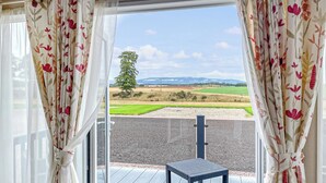 Una habitación con vista a un campo verde y montañas, una mesita y cortinas con estampado floral.