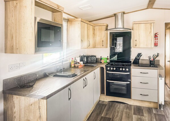 A compact kitchen with wooden cabinets, a microwave, a stove, and a sink. - Modern and contemporary 12ft wide caravan with one double and one twin. (Bleadon, Weston-Super-Mare)