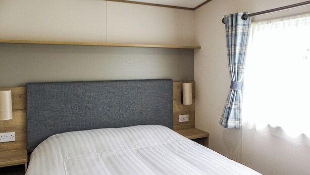 Een enkel bed met een grijs hoofdbord, bedlampjes en een raam met gordijn.