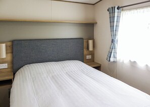 A single bed with a grey headboard, bedside lamps, and a window with a curtain. - Modern and contemporary 12ft wide caravan with one double and one twin. (Bleadon, Weston-Super-Mare)