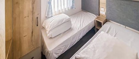 Un lit simple avec une tête de lit grise, une armoire en bois avec deux tiroirs et une fenêtre avec des rideaux.