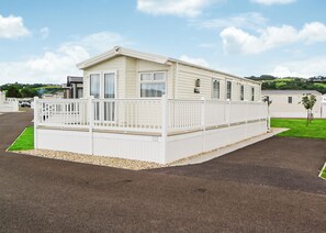 A single-story mobile home with a fenced deck, a driveway, and a grassy area in the background. - Modern and contemporary 12ft wide caravan with one double and one twin. (Bleadon, Weston-Super-Mare)