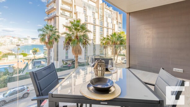 Uma varanda com mesa de vidro, cadeiras e vista para um edifício com varandas e palmeiras.