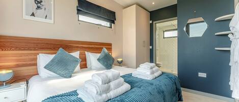 A modern bedroom with a wooden headboard, a large bed with blue bedding, a bedside table with a lamp, and a shower area.