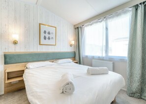 Una cama bien tendida con sábanas blancas y una toalla enrollada, un cuadro en la pared, y una ventana con cortinas.