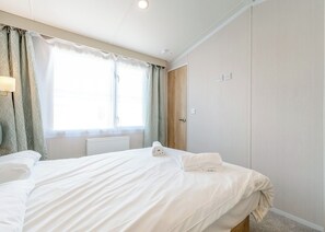 A hotel room with a bed, a radiator, a wooden wardrobe, and a window with curtains. - Platinum Caravan 6 (Pet) (Skegness)