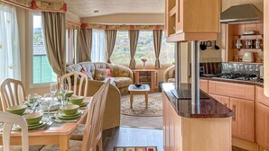 Une pièce chaleureuse avec une table à manger dressée pour quatre personnes, un canapé, un coin cuisine avec une cuisinière et de grandes fenêtres avec des rideaux.