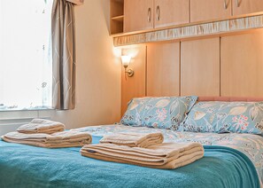 A hotel room with a bed, pillows, a nightstand, a lamp, and a window with curtains. - Traditional yet comfortable caravan with one double and two twins. (Dolgellau)
