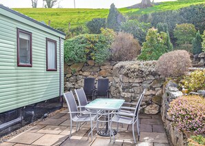 A patio with a table and chairs, a stone wall, and a green building. - Traditional yet comfortable caravan with one double and two twins. (Dolgellau)
