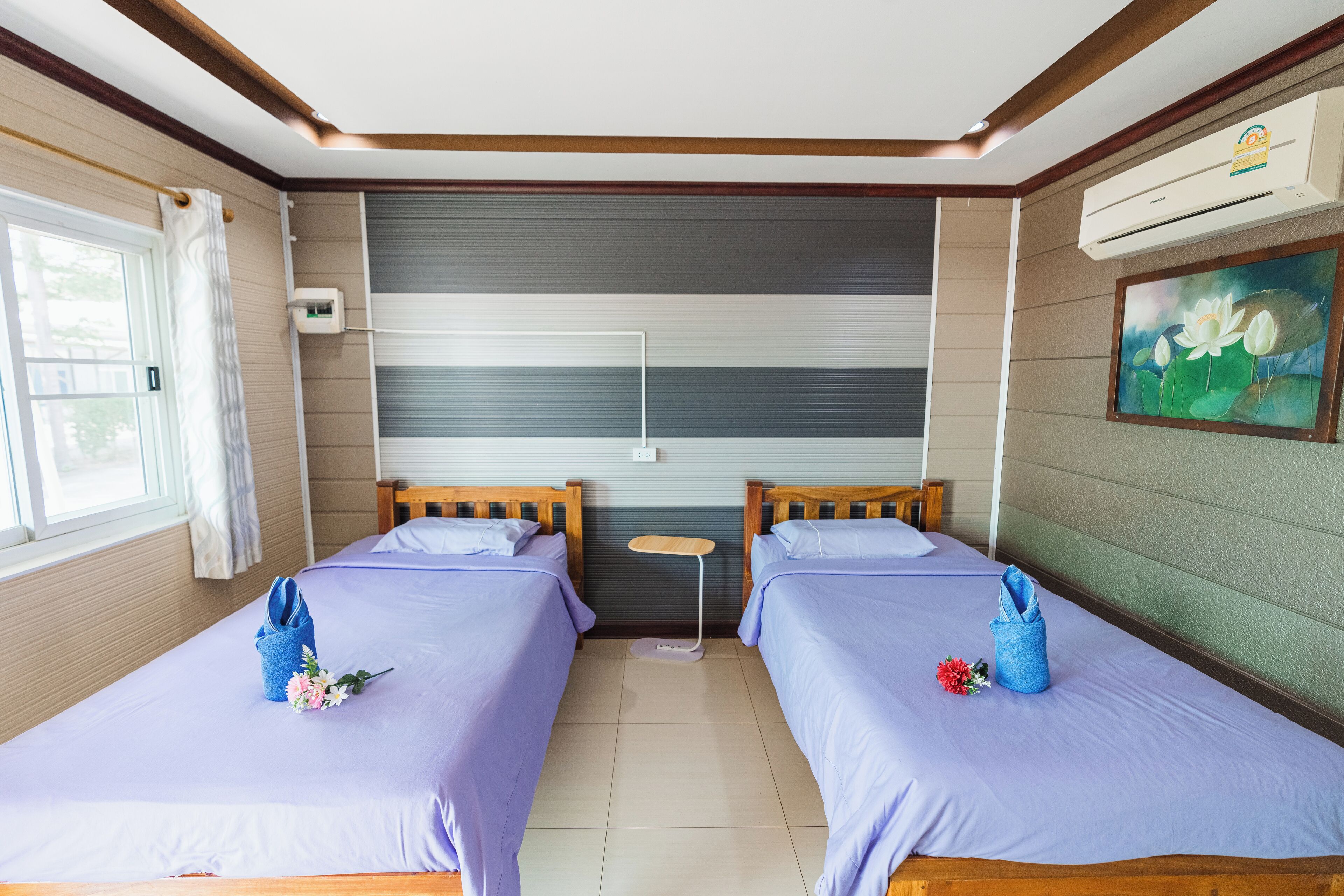 A hotel room with two beds, a small table, an air conditioning unit, and a painting on the wall.
