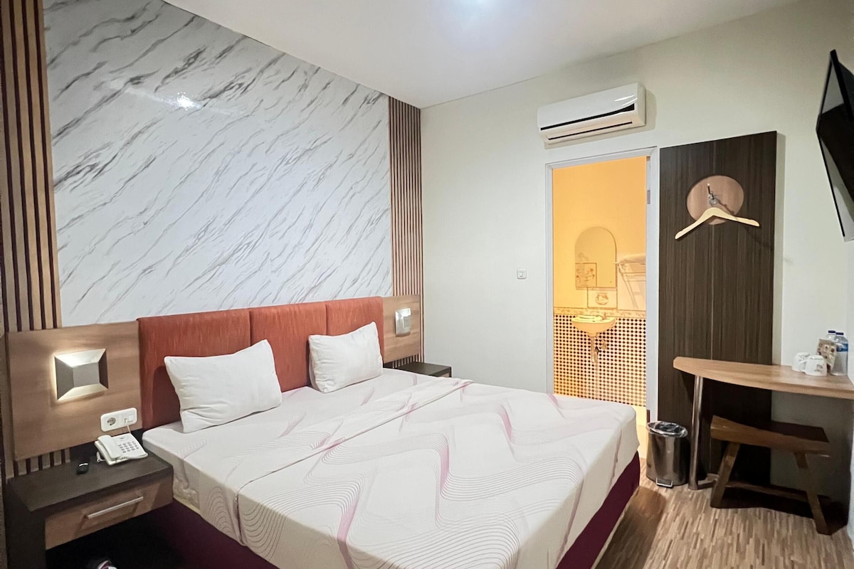 A hotel room with a large bed, bedside tables, a desk, and an air conditioning unit.