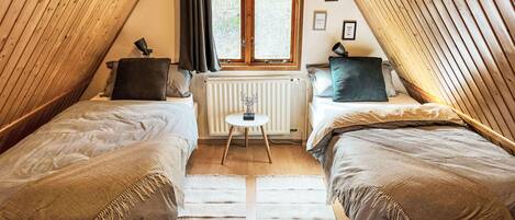 A cozy attic bedroom with two single beds, a small table, and a window with curtains.