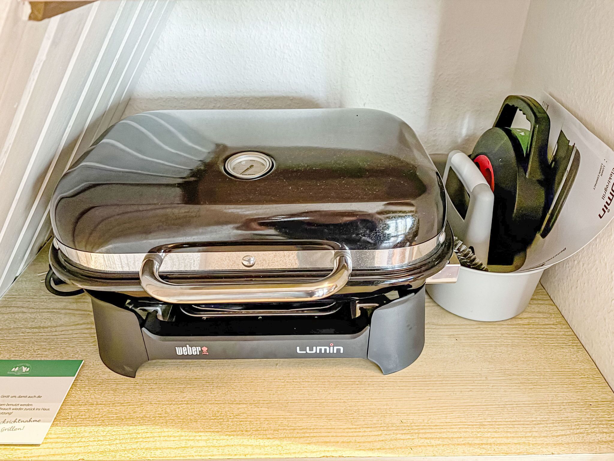 Una parrilla portátil Weber Lumin con tapa, sobre una superficie de madera junto a un recipiente blanco que contiene utensilios para asar.
