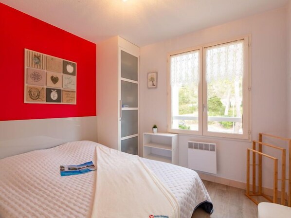 A bedroom with a bed, a red accent wall, a window with a view, and a shelf with decorative items. - Les Coraux by Interhome (Agay Saint Raphael)