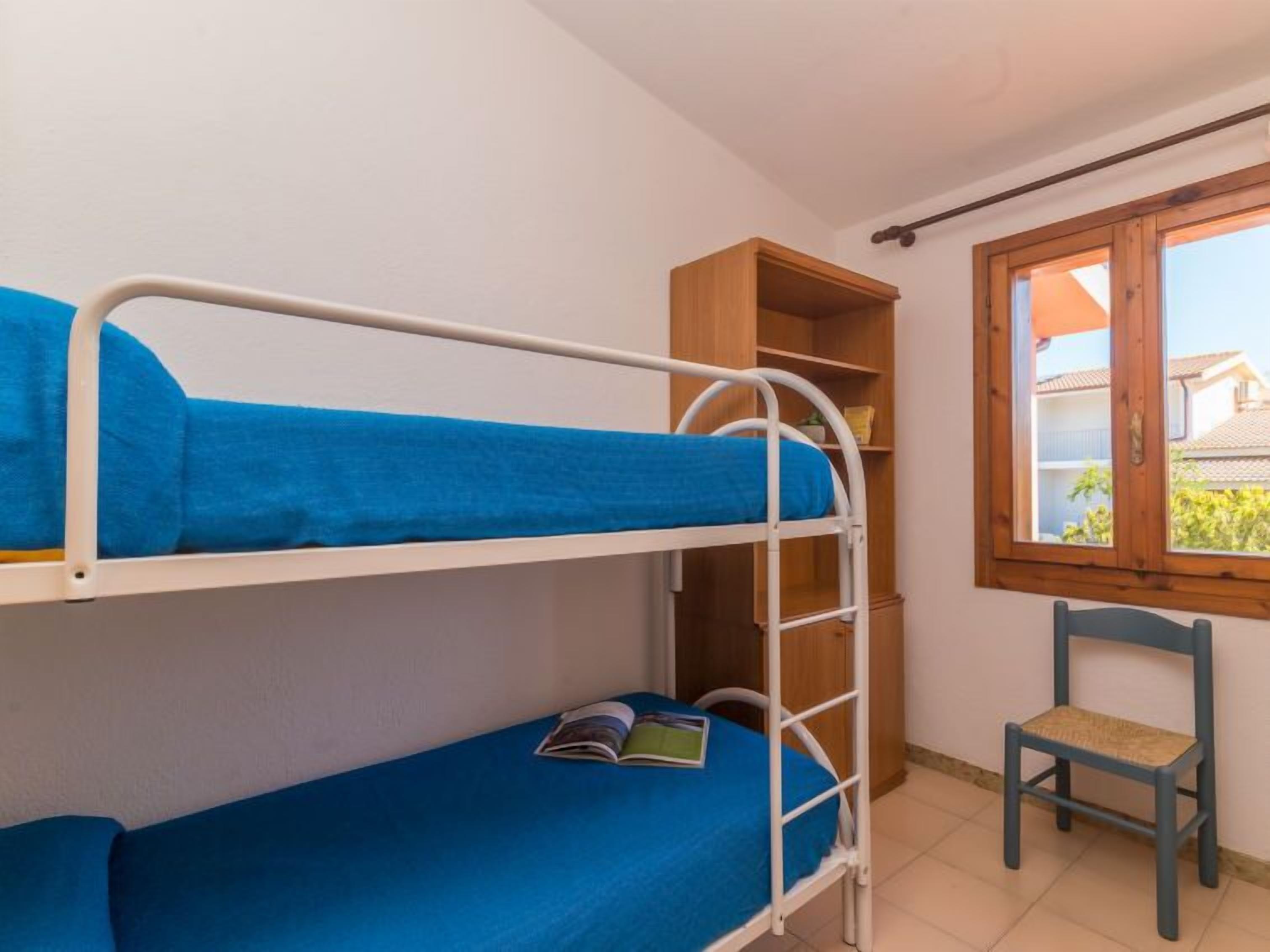 Uma cama beliche com roupa de cama azul, um guarda-roupa de madeira, uma cadeira e uma janela com vista para edifícios.
