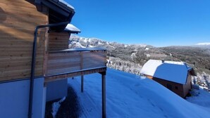 Une cabane en bois avec un escalier en métal menant à une terrasse, d’où l’on peut admirer un paysage montagneux enneigé.