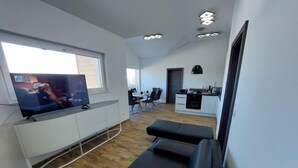 Un salon moderne avec un téléviseur à écran plat, un canapé en cuir noir, un plancher en bois et un coin cuisine à l’arrière-plan.