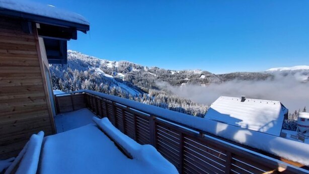 Un balcon en bois, avec des balustrades recouvertes de neige, d’où l’on peut admirer des montagnes et des arbres également recouverts de neige.